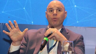 Jay Heller fala durante painel, usando terno azul e microfone, em frente a um fundo azul com elementos gráficos