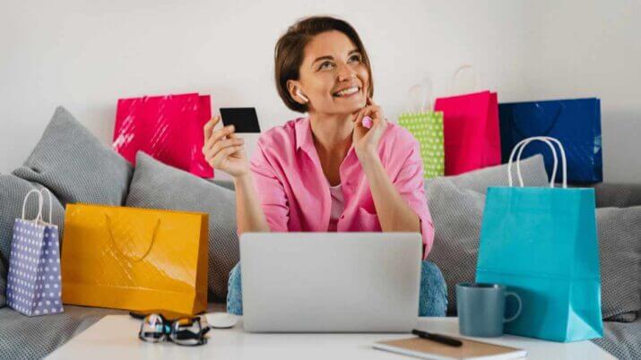Mulher segura cartão de crédito enquanto faz compras na internet, rodeada de sacolas coloridas