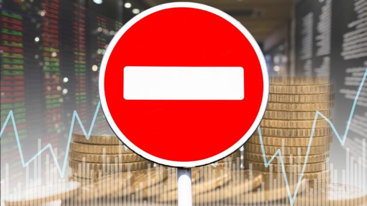 Montagem com um gráfico de ações ao fundo, uma pilha de moedas na parte inferior e, em primeiro plano, uma placa vermelha de proibido