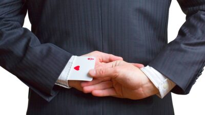 Imagem mostra um homem de terno azul escuro, de costas, tirando uma carta de As da manga esquerda