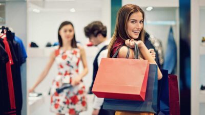 Mulher carrega sacolas de compras em uma loja em um shopping. Atrás dela estãou outros consumidores