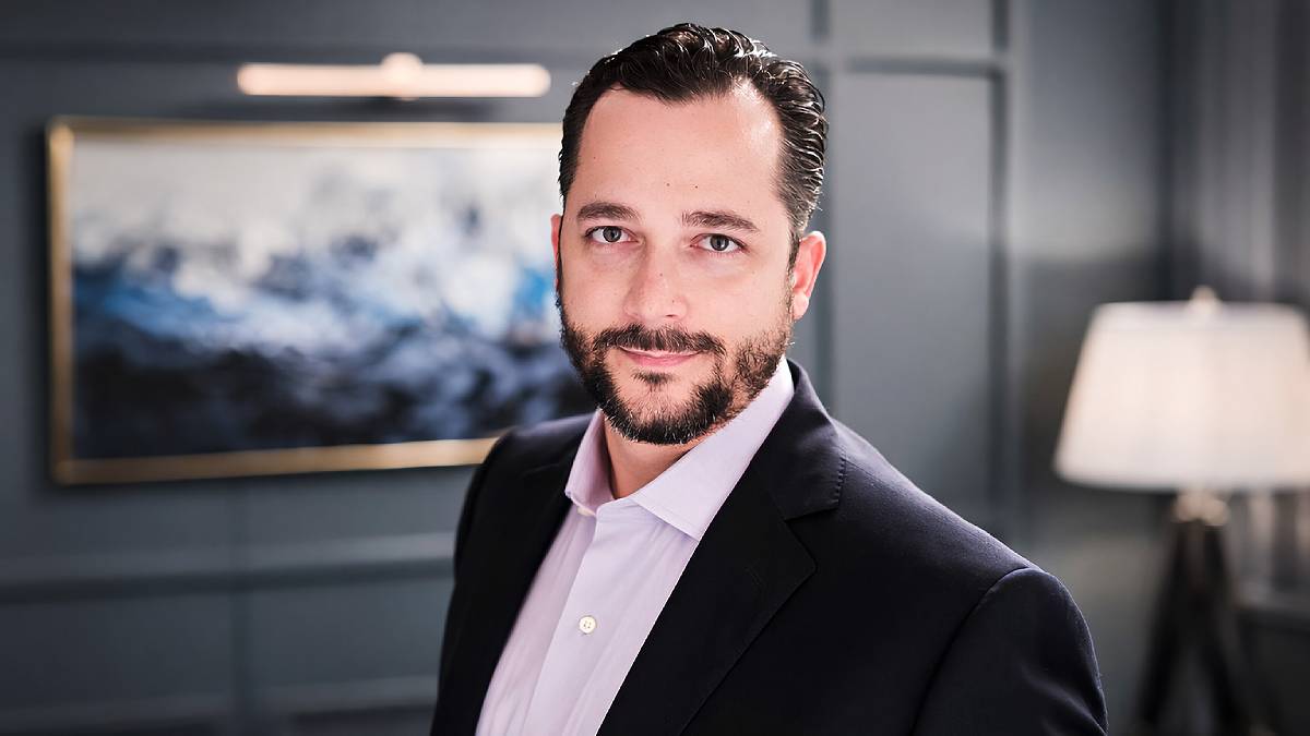Foto do CIO do EFG Private Wealth Management, Luis Ferreira, em uma sala com uma quadro ao fundo. Ele usa terno azul escuro e camisa rosa claro.