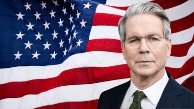 Montagem com a bandeira dos EUA ao fundo e, em primeiro plano, o secretário do Tesouro dos EUA, Scott Bessent. Ele é grisalho, usa óculos e veste terno azul escuro, com camisa branca.