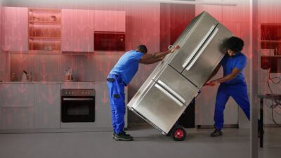 Carregadores com uma geladeira e, ao fundo, uma ilustração de gráfico em queda para representar a desvalorização das ações da Whirlpool (WHR), dona da Brastemp