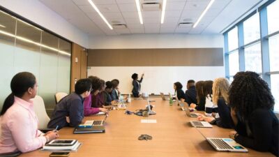 Pessoas em volta de uma mesa de reunião com notebooks na frente e uma mulher de pé próxima a um quadro branco