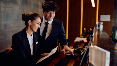 Happy receptionists cooperating while working on a computer at hotel front desk.