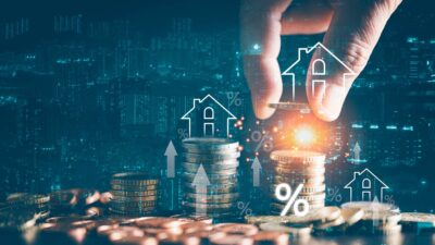 Mão colocando uma moeda em cima de pilha de moedas com ilustrações de casas e setas para cima, representando valorização de investimentos imobiliários