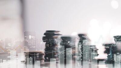 Vista da cidade em segundo plano, com pilha de moedas e gráficos de investimentos para representar ativos imobiliários na bolsa de valores.