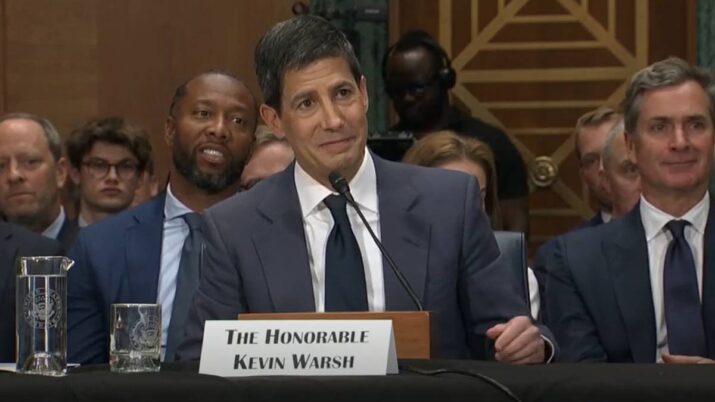 Frame do YouTube com Kevin Warsh sendo sabatinado no Senado dos EUA. Ele está sentado, com um microfone à frente. Veste terno azul matinho e camisa branca.