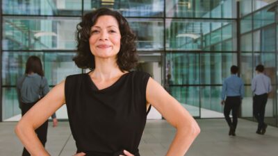 Mulher com camiseta preta coloca as mãos na cintura e faz uma pose confiante. Ela está em frente a um prédio corporativo, com outros colegas indo em direção à porta de entrada ao fundo.