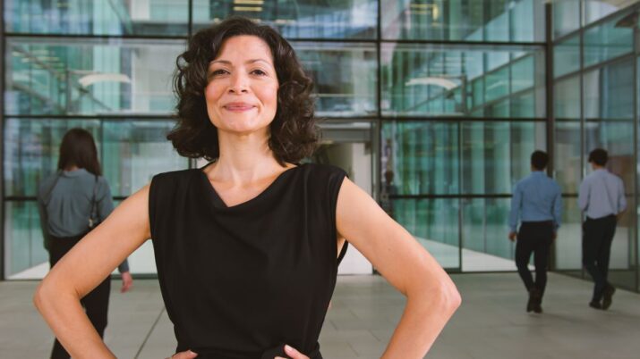 Mulher com camiseta preta coloca as mãos na cintura e faz uma pose confiante. Ela está em frente a um prédio corporativo, com outros colegas indo em direção à porta de entrada ao fundo.