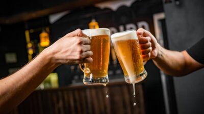 Imagem mostra duas mãos brindando com canecas de chope em um bar ou restaurante