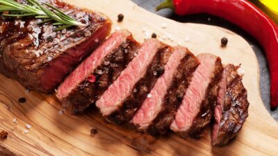 Imagem mostra uma peça de carne ao ponto, cortada sobre uma tábua de madeira, com temperos ao redor