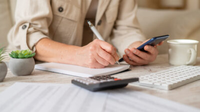 Woman’s hands writing notes in a spiral notebook while holding a smartphone, actively managing personal finances, calculating a budget, and planning household expenses on a desk
