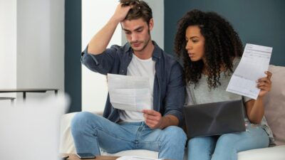 Imagem mostra um casal sentado no sofá de casa com contas a pagar. Eles parecem preocupados com dívidas ou inadimplência.