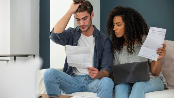 Imagem mostra um casal sentado no sofá de casa com contas a pagar. Eles parecem preocupados com dívidas ou inadimplência.