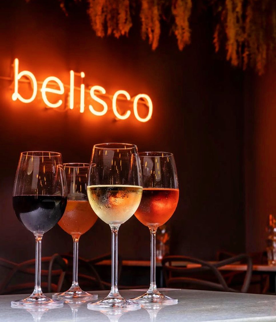 quatro taças de vinho em cima de uma mesa com letreiro vermelho escrito "belisco" atrás