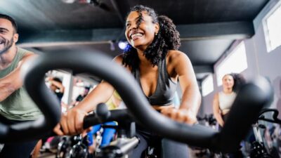 Imagem mostra uma mulher com roupa de ginástica em uma aula de spinning em uma academia,