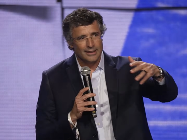 André esteves, de terno e óculos, com microfone na mão esquerda, em cima do palco