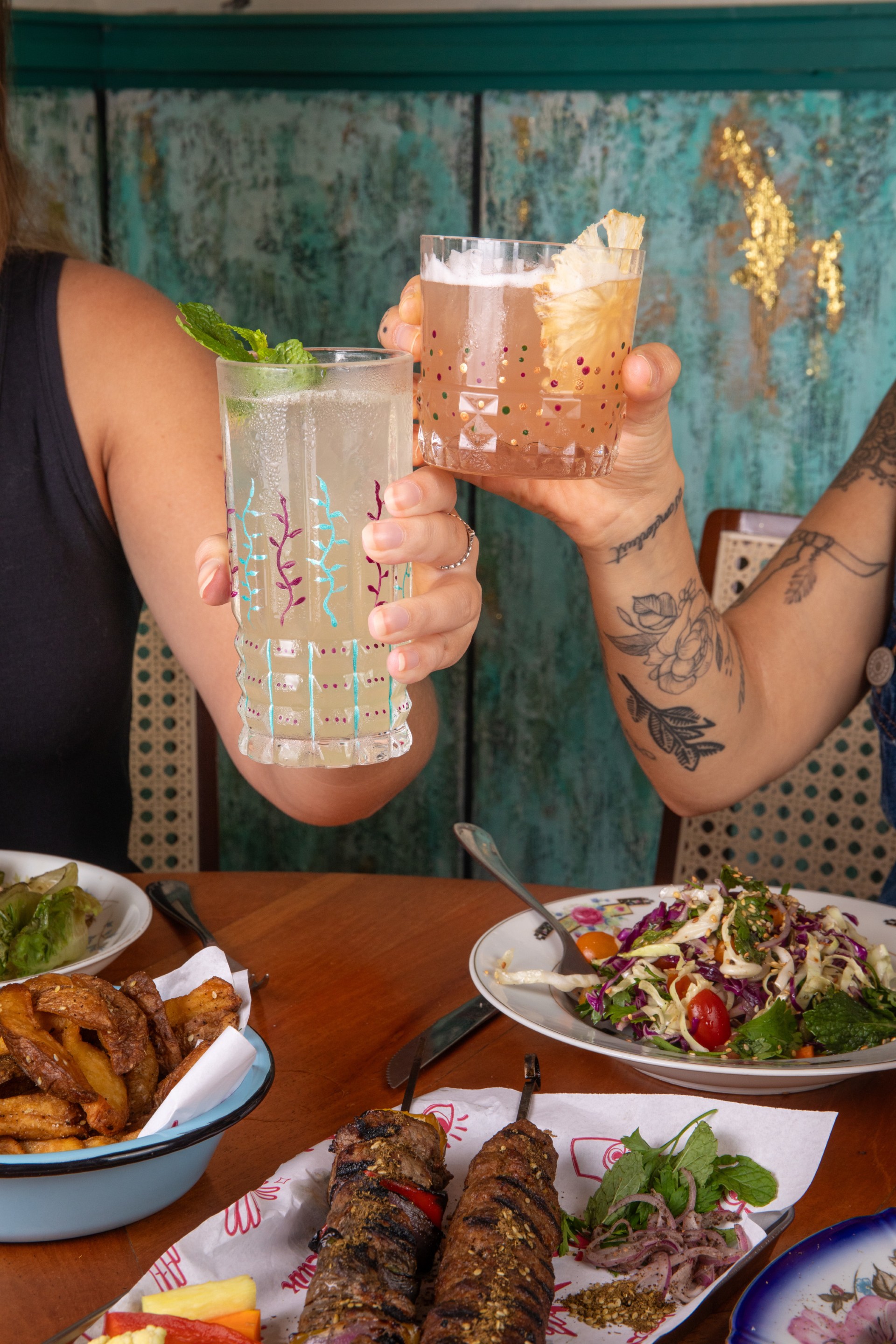 duas pessoas fazendo um brinde com drinks e comida árabe na mesa