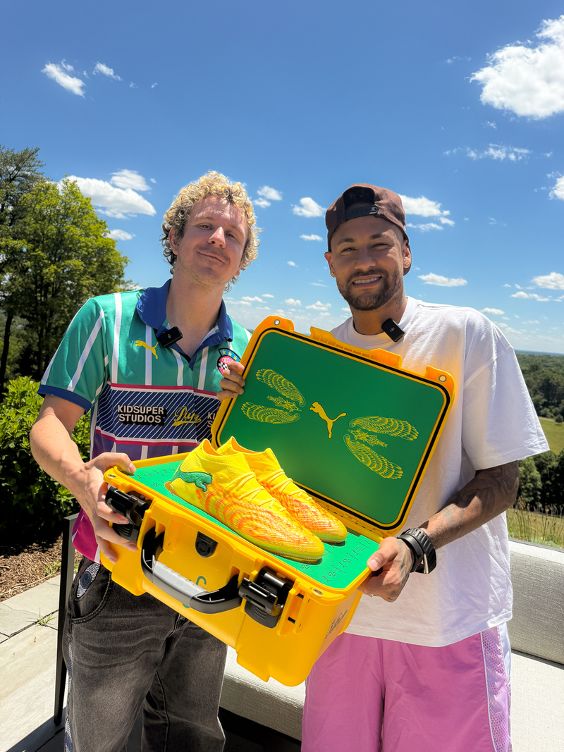 Neymar Jr à esquerda e Colm Dillane à direita, ambos segurando uma maleta amarela aberta com a chuteira à mostra