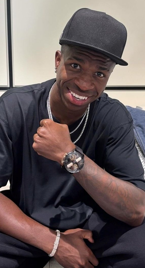vini sentado sorrindo com boné e roupas pretas mostrando o relógio no pulso