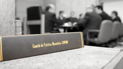 homens reunidos em uma mesa em segundo plano, com um caderno escrito Comitê de Política Monetária (Copom) em primeiro plano.