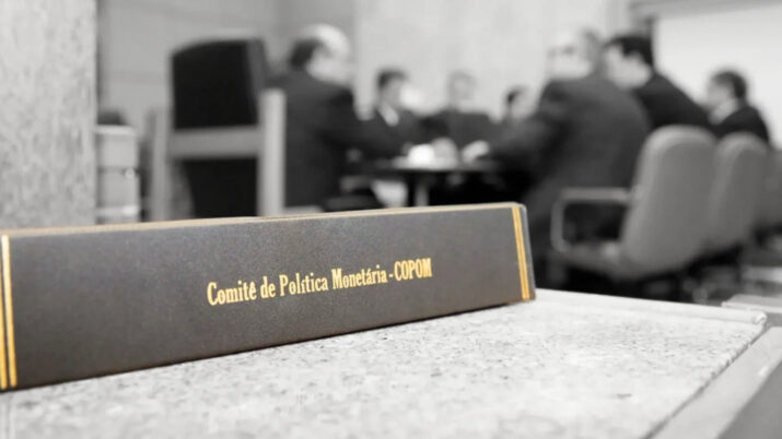 homens reunidos em uma mesa em segundo plano, com um caderno escrito Comitê de Política Monetária (Copom) em primeiro plano.