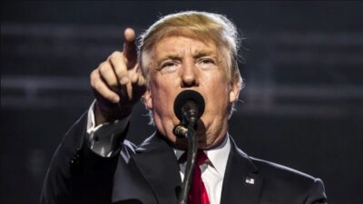 Donald Trump está de pé, em frente a um microfone, apontando com do dedo para frente. Ele usa terno azul marinho, camisa branca e gravata vermelha.