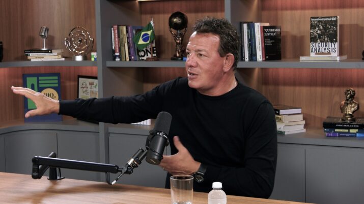 Com um estante ao fundo, um fome de blusa preta está sentado em uma mesa, com um microfone a frente