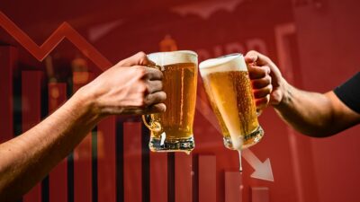 Imagem mostra duas mãos brindando com canecas de chope em um bar ou restaurante