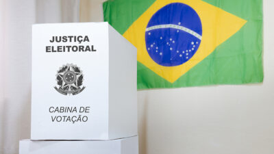 Bandeira do Brasil pendurada em uma parede ao fundo da imagem. Em primeiro plano, a cabine de votação das eleições no Brasil.
