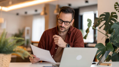 Homem sério da geração millennial usando laptop sentado à mesa em um escritório em casa, sujeito concentrado em roupas casuais olhando para o jornal, comunicando-se online, escrevendo e-mails, trabalhando ou estudando remotamente no computador em casa.