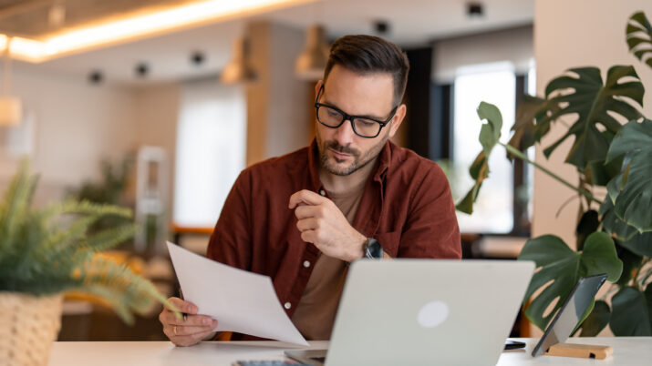 Homem sério da geração millennial usando laptop sentado à mesa em um escritório em casa, sujeito concentrado em roupas casuais olhando para o jornal, comunicando-se online, escrevendo e-mails, trabalhando ou estudando remotamente no computador em casa.