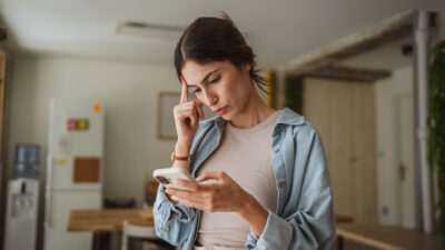 A jovem empresária está com dificuldades enquanto trabalha em casa e lê uma mensagem no celular, tocando a testa com uma expressão pensativa.