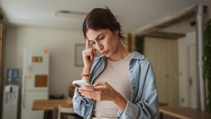 A jovem empresária está com dificuldades enquanto trabalha em casa e lê uma mensagem no celular, tocando a testa com uma expressão pensativa.