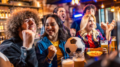 Amigos comemorando um gol com uma bola de futebol e batatas fritas no balcão do bar. Grupo de torcedores assistindo a uma partida de futebol juntos no interior rústico de um pub.