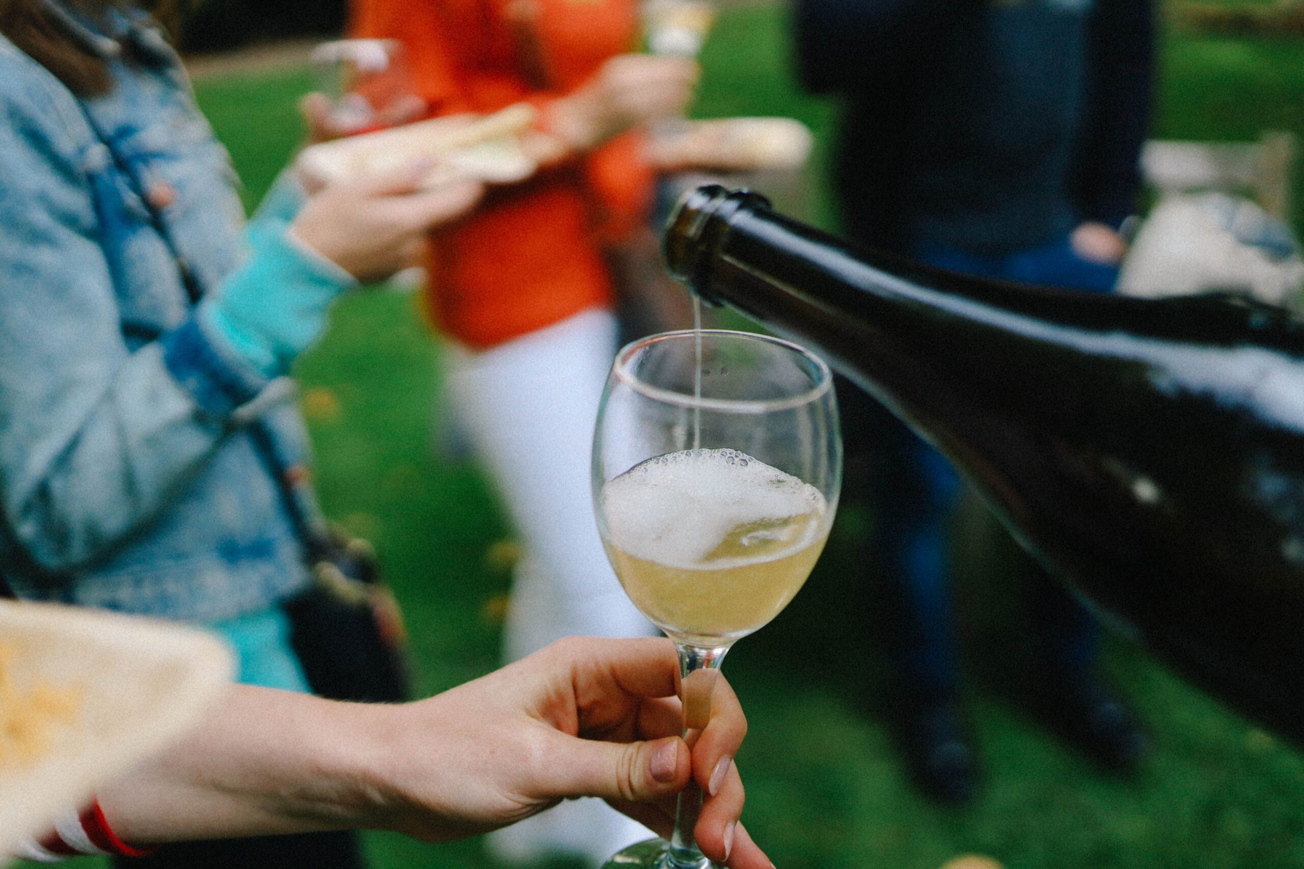 Mão estende taça de vinho branco e alguém lhe serve um espumante