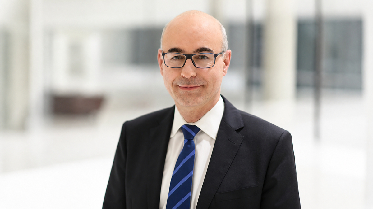 Achim Wambach, presidente do instituto de pesquisa econômica ZEW, aparece em primeiro plano. Ele usa óculos, é calvo e está de terno preto, camisa branca e gravata listrada de tons de azul.