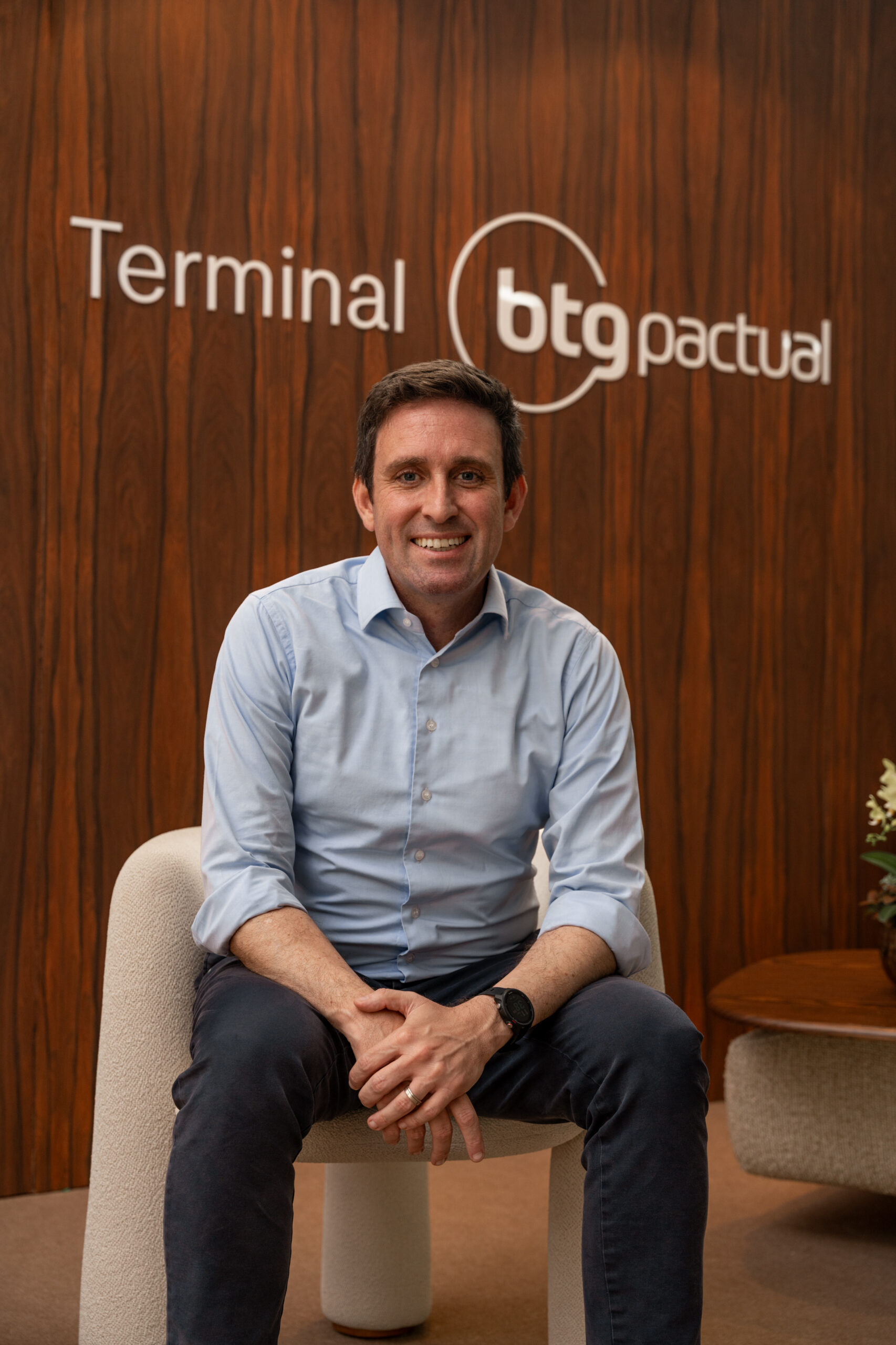 Fabio Camargo, o CEO do Terminal BTG Pactual, aparece sentado em uma poltrona branca, usando calça azul escuta, camisa azul clara e um relógio. Ele está sorrindo. Atrás dele, em uma parede revestida de madeira lê-se "Terminal BTG Pactual"