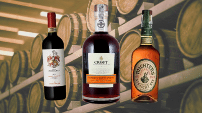 Três garrafas de vinho, vinho fortificado e whisky; ao fundo, barris de madeira