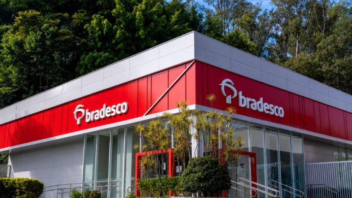 Fachada de uma agência do Bradesco com arquitetura moderna, destacando painéis vermelhos e brancos, logotipos do banco em ambas as laterais do prédio, amplas paredes de vidro na entrada, corrimãos de acessibilidade e paisagismo com árvores e arbustos ao redor.