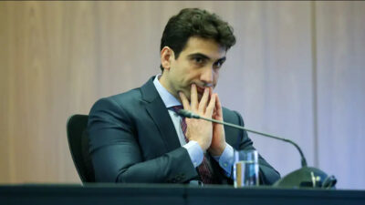 Gabriel Galípolo, presidente do Banco Central do Brasil, está sentado, com um microfone e um copo de água sobre a mesa que está a sua frente