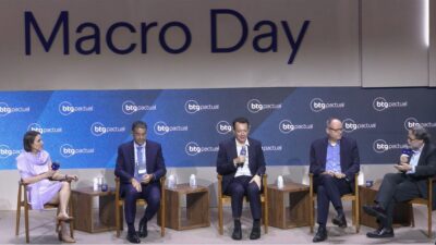 Quatro economistas do BTG Pactual estão sentados em cadeira de madeira. Todos eles vestem terno azul. A mediadora está de lilás. Ao fundo, um painel com o logo Macro Day do BTG Pactual.