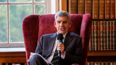 O economista Mohamed El-Erian está sentado em uma poltrona vermelha, segurando um microfone. Atrás dele, uma estante de livros.