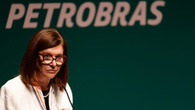 A presidente da Petrobras, Magda Chambriard, está de pé, em frente a um microfone. Atrás dela, um painel verde com o nome da Petrobras em branco.