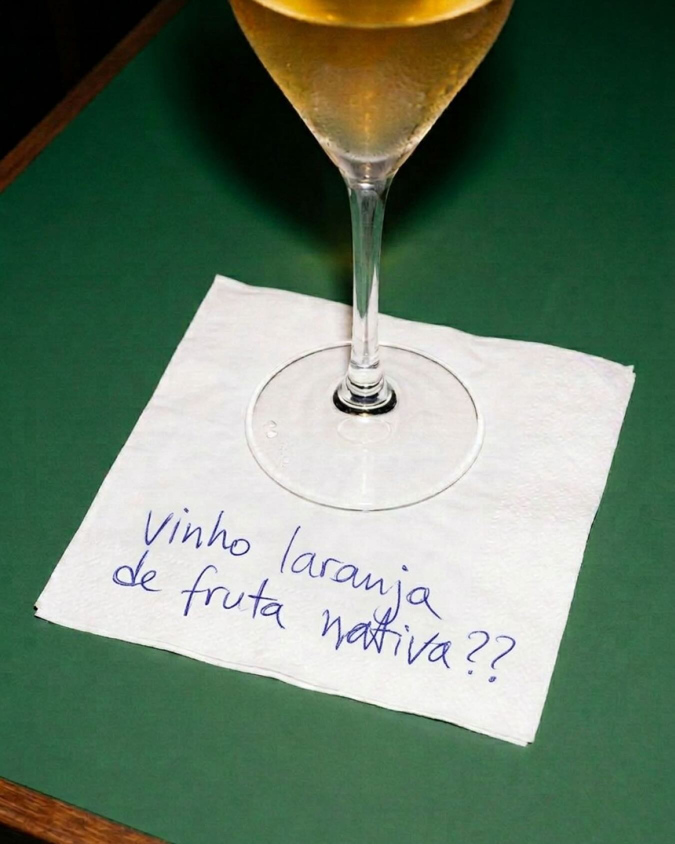 Taça com bebida dourada sobre um guardanapo, onde se lê: "vinho laranja ou fruta nativa??"