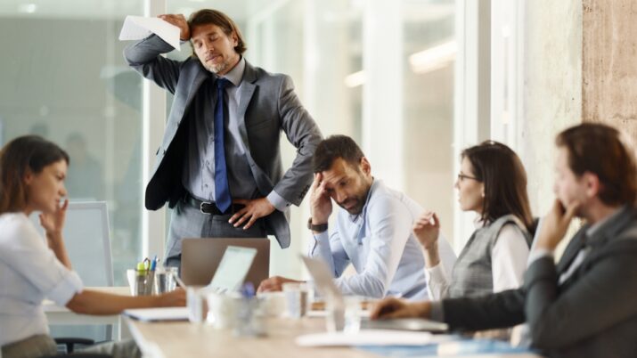 Imagem mostra uma equipe de pessoas em ambiente de trabalho, em uma sala de reunião de escritório, vestindo roupas formais. Eles estão discutindo e com expressões de preocupação no rosto