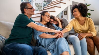 Imagem mostra pessoas de uma família em um sofá, sendo um homem e uma mulher mais velhos e duas mulheres mais jovens. Eles estão se consolando, tristes, em luto ou preocupados.