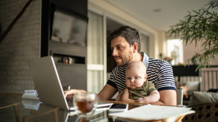 Homem segurando seu filho recém-nascido e trabalhando com laptop em casa